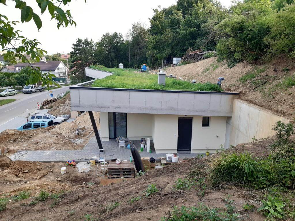 Rozestavěný podzemní dům ve svahu, pohled zdola: světlá fasáda, terasa, betonová stěna u navezené zeminy, zelená střecha.