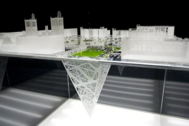 White scale model of a historic city block with green plaza, an inverted lattice cone descending into darkness below