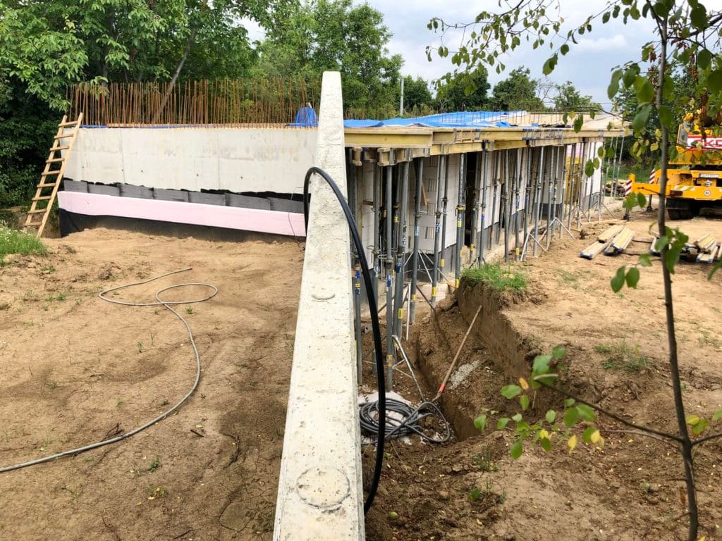 Earth-sheltered house shell under construction, concrete roof slab on shoring props above window openings, insulation layers exposed, crane in background