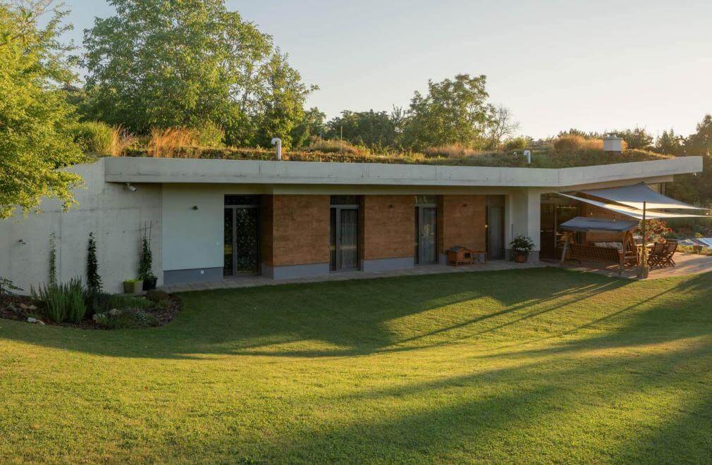 Earth-sheltered home with wildflower roof, wood-clad facade and pool-side sail shade at sunset