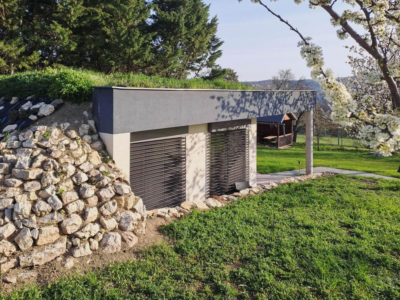 Podzemný dom Balaton zapustený do svahu s trávnatou strechou, zatvorené okenice, kamenný múr a terasa smerom k lúke.
