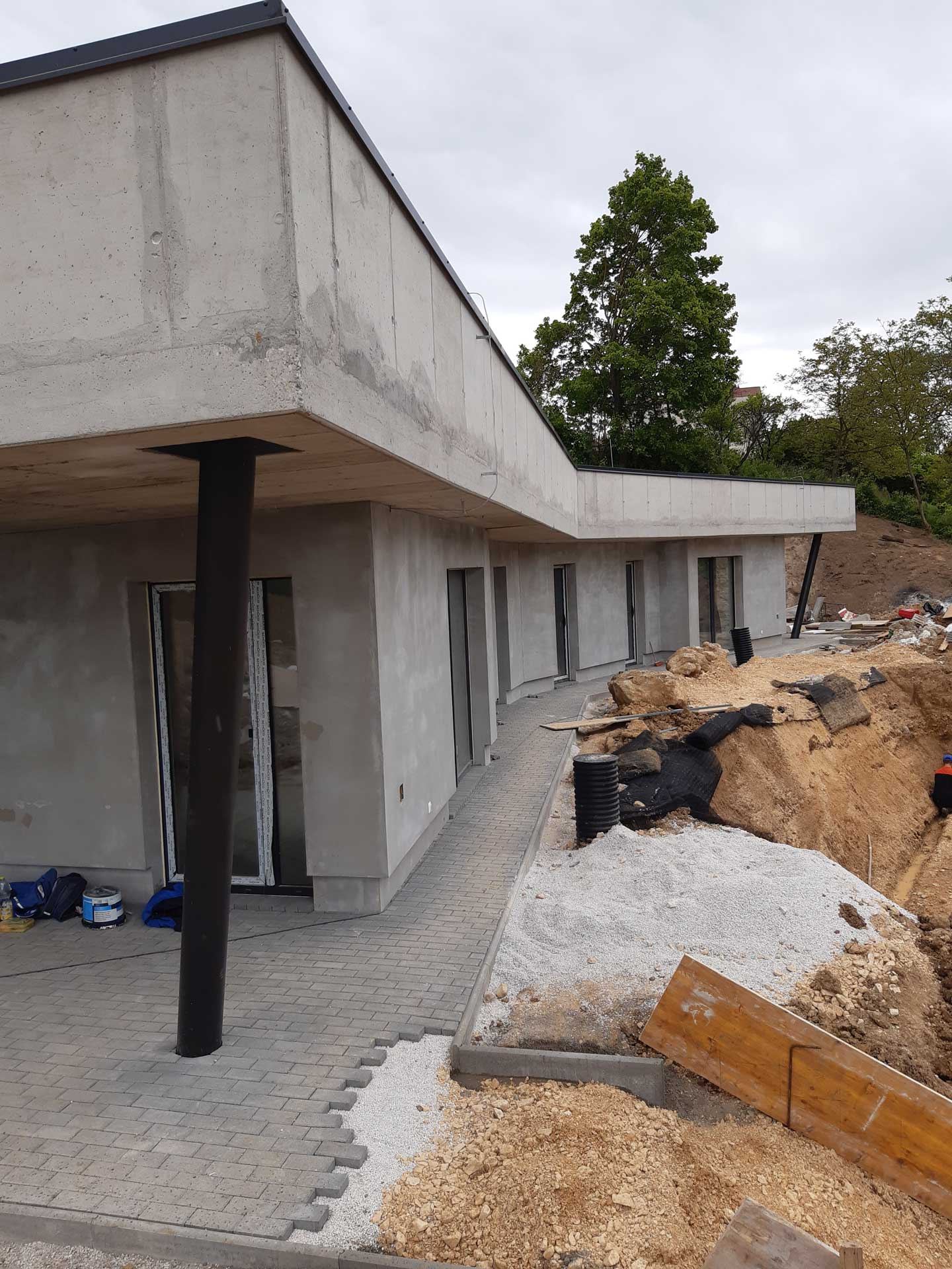 Podzemný dom v Trenčíne vo výstavbe, presklená fasáda na tenkom stĺpe, terasa vedľa rozkopanej zeminy s rúrami