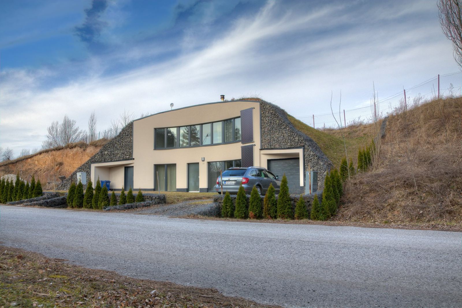 Earth-sheltered house in Rohoznik with cream front wall between grassy gabion-walled mounds and a parked car