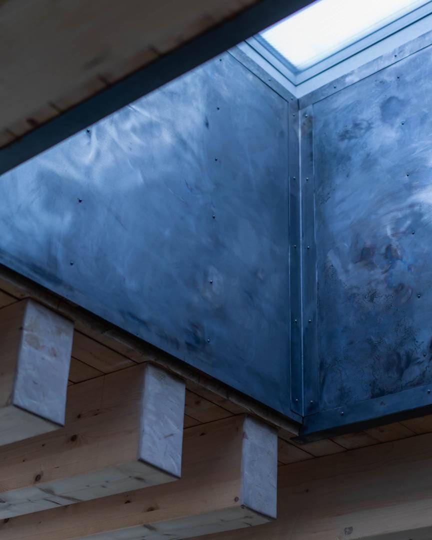 Pohled zdola na světlík podzemního domu, sešroubované kovové panely nad odhalenými dřevěnými trámy.